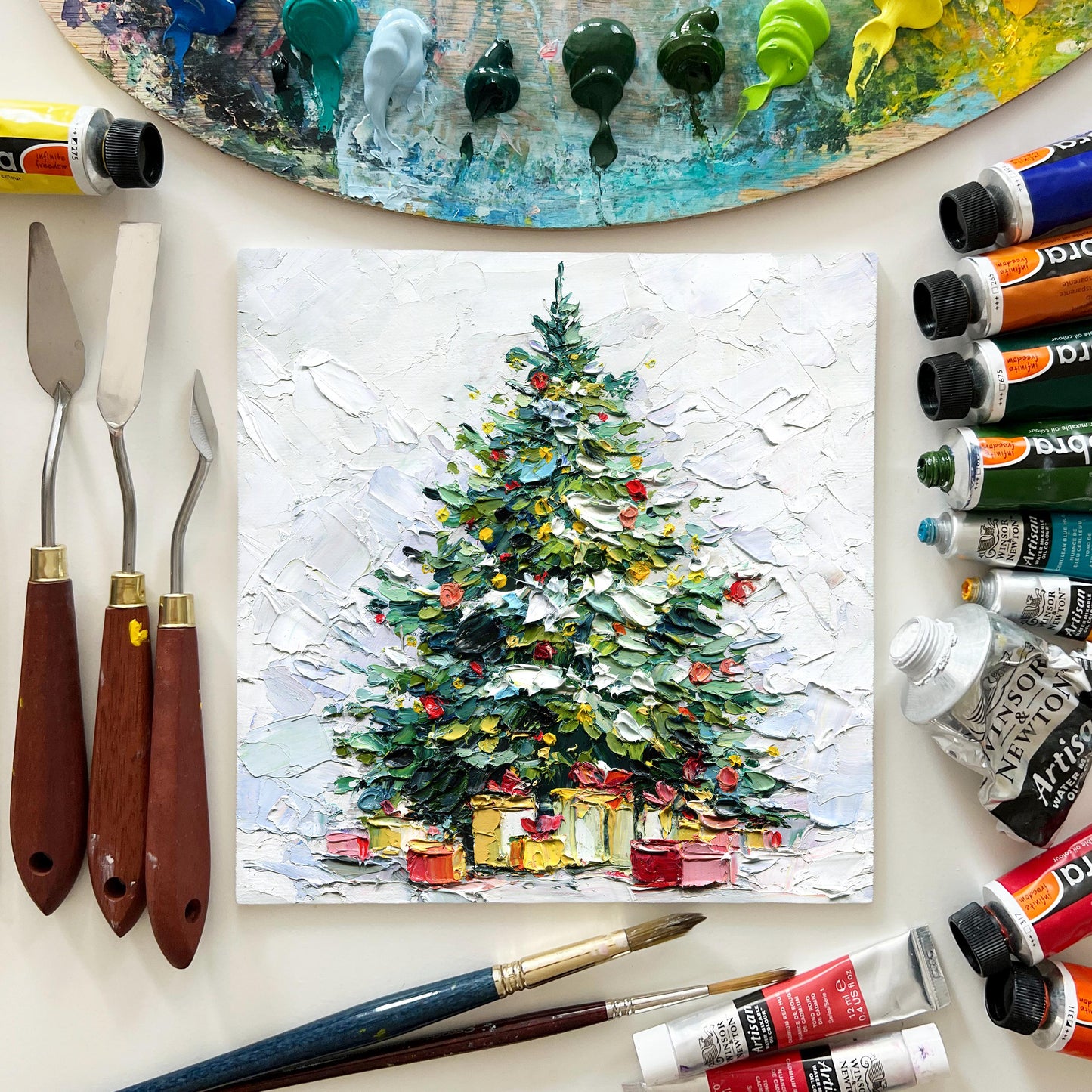 Painting of a Christmas tree with presents, surrounded by art supplies including paint tubes and brushes.