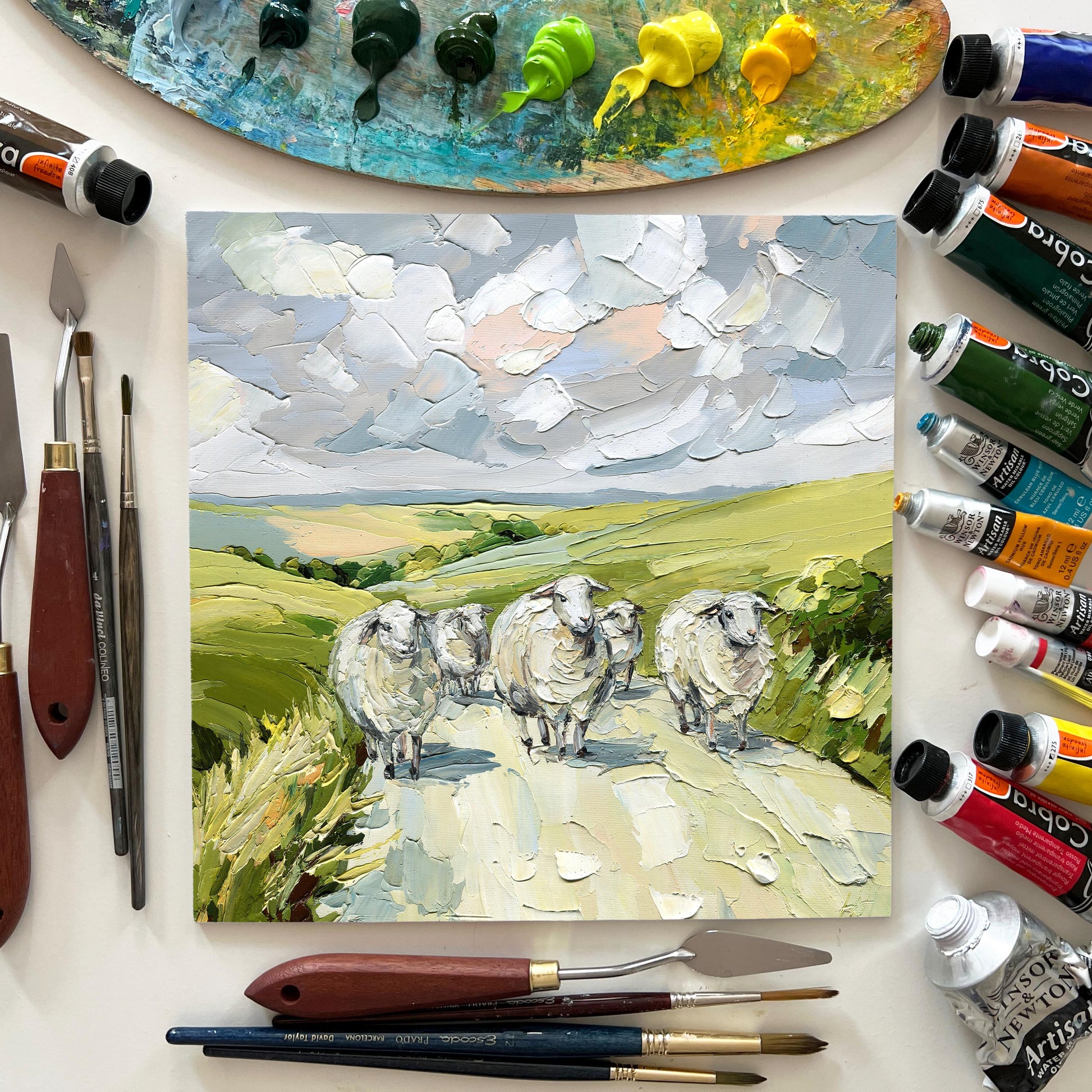 Painting of sheep on a landscape with art supplies including paint tubes and brushes.