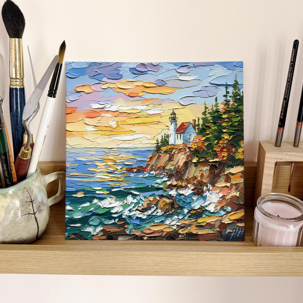Unframed Acadia Lighthouse painting on a shelf beside brushes and a candle
